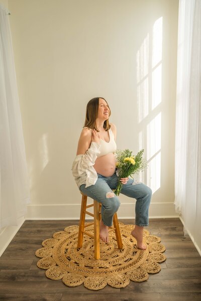 mom sitting on wood stool on a jute rug holding yellow and white flowers white looking out the window and laughing