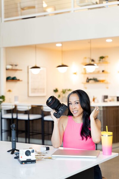 Creative headshots of a Dallas brand photographer dressed in a pink t-shirt, holding her professional camera and surrounded by tools of her trade in a studio setting.