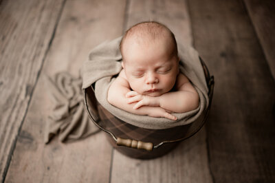 Newborn with hands folded under chin in brown bucket