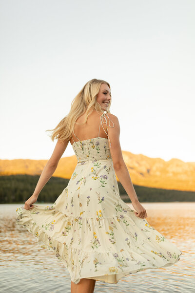 A young woman smiling while twirling  in front of a lake and mountains.