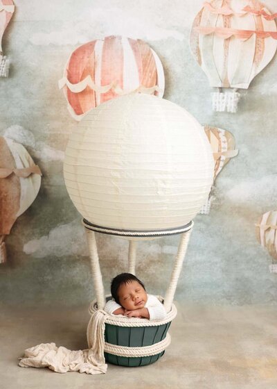 A charming scene with a newborn baby cozy in a basket that looks like a hot air balloon. The background features soft, muted balloons, creating a dreamy and whimsical feel. The baby is peacefully resting, adding warmth to the playful setup.