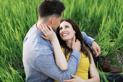 Two lovers looking at each other and smiling brightly as they sit in tall grass at Elm Creek Park Reserve. The man has his arms around her as she gently touches his head. Captured by Toly Dzyuba Photography in Minnesota.