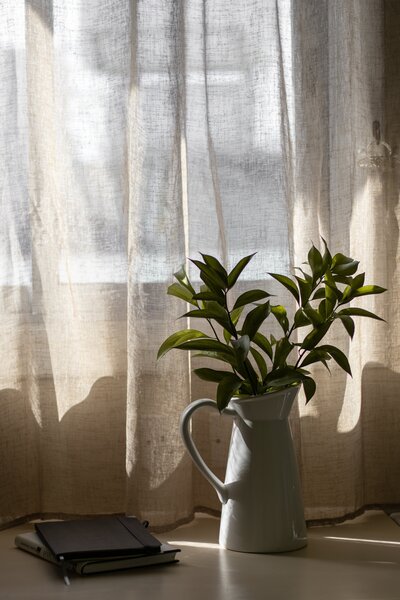 Vase with greenery next to notebooks and a window with a linen curtain