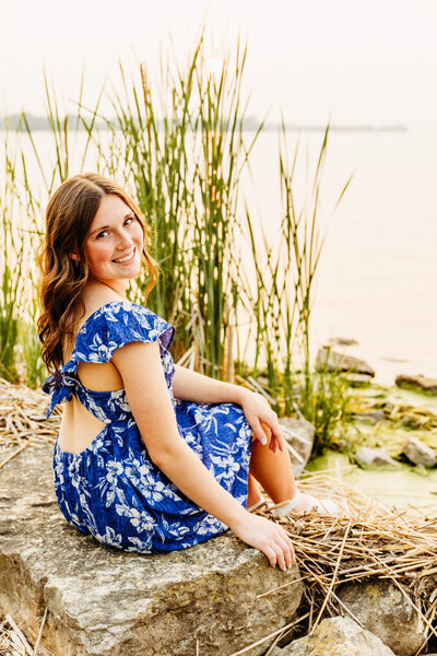 teen girl in a blue and white dress sitting on a rock near a lake