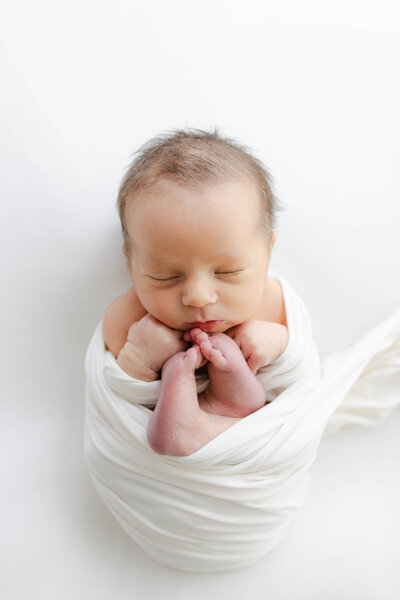 baby swaddled in white with his hands and feet by his face, Indianapolis newborn photogrpaher
