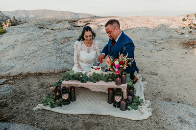 A couple enjoys a romantic Lake Tahoe elopement, sitting on a blanket by the sandy shore, toasting drinks as the sun sets. The woman wears a blue and white striped dress, and the man wears a white shirt. Snow-capped mountains and the lake’s clear waters create a stunning backdrop.