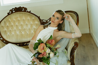 A bride in a simple, sleek white wedding dress is sitting on a vintage and elegant gold couch. She is almost lying down and has one arm propped on the arm of the couch, gently touching her head.