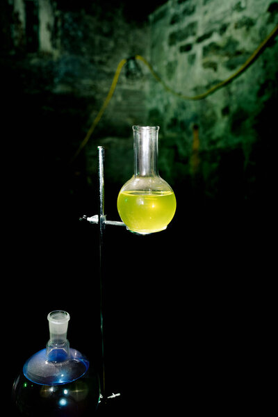 A laboratory setup with a round-bottom flask containing a yellow liquid, suspended on a metal stand against a dark, rustic stone wall background.
