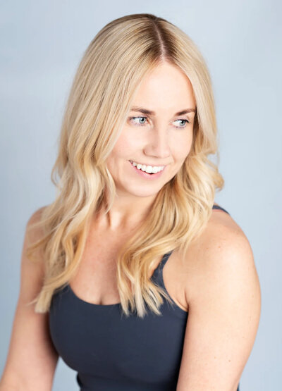 Confident yoga practitioner Jen Carwardine smiles warmly, her blonde hair styled in waves, embodying the positive impact of creative sequencing in her yoga practice, against a soft grey background.
