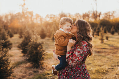 Golden hour photo of mother holding and kissing little boy on Christmas tree farm