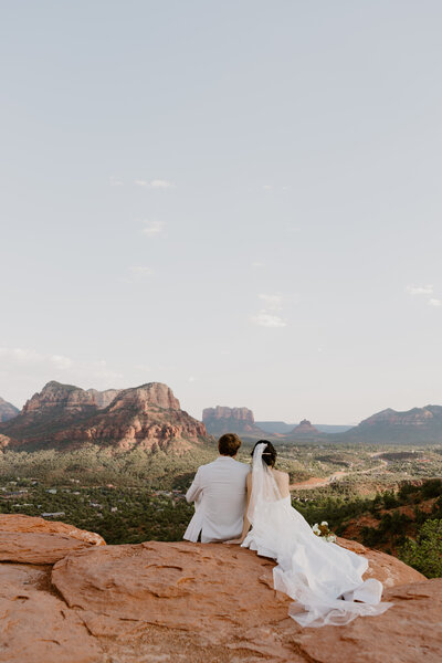 Wedding couple leans on Sedona ledge with backs to camera.

