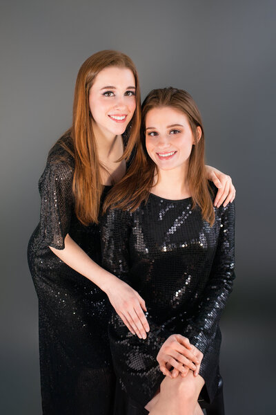 Two sisters with red hair wear black dresses and embrace one another in front of a gray backdrop