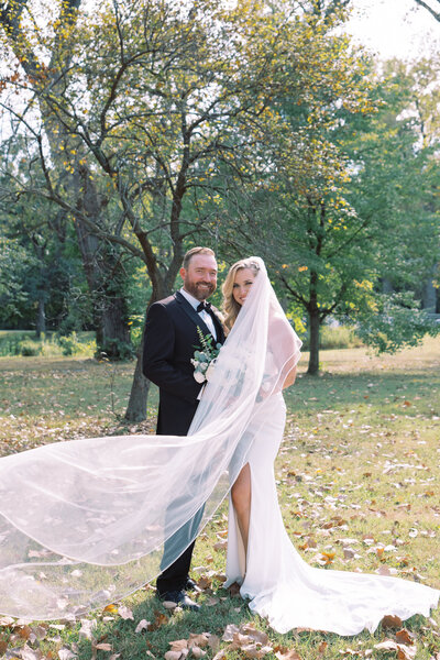 film inspired photo of a traditional bride and groom pose in a lush green park with the veil blowing in the wind