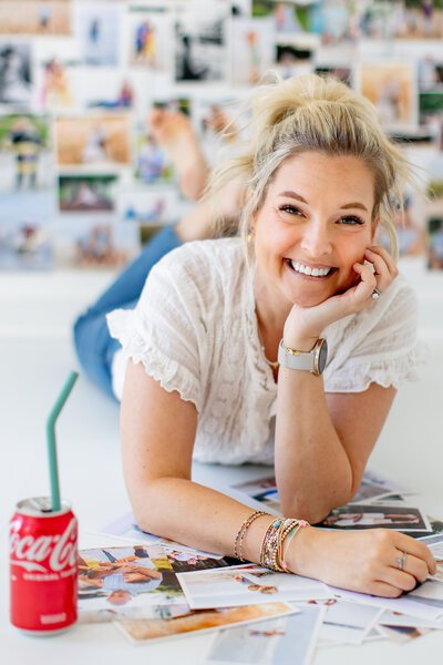 Brooke, a Virginia Beach family photographer, smiling while lying among printed portraits of families she has captured.  A Coca-Cola can sits nearby, adding a nostalgic touch.  Behind her, a collage of family photography moments reflects her storytelling approach.