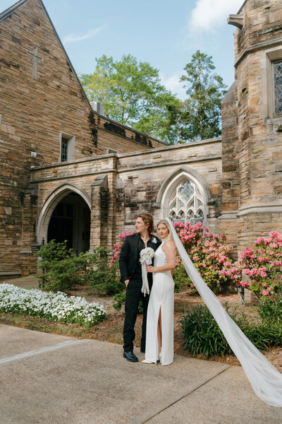 Bride and groom posing in front of a historic stone church with bride’s veil flowing — elegant Alabama wedding portrait by EC Photography