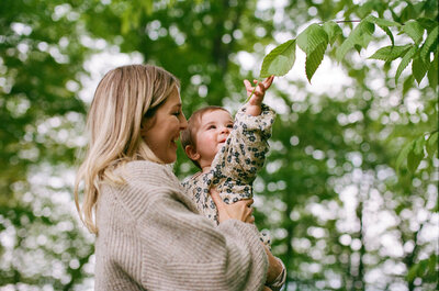 A smiling mother holds her daughter up next to a tree in a film photography session by Oshawa family film photographer, Jennifer van Son.
