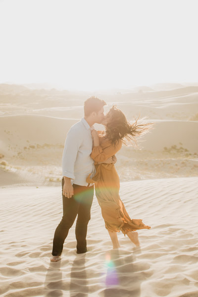 jackson hole photographers  captures engagement photos in a sand dune with man and woman kissing as the sun sets right over their heads and the wind blows the woman orange dress and hair
