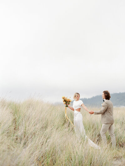 bride holding hands with groom and leading him walking away in a tall field of seagrass at the beacj