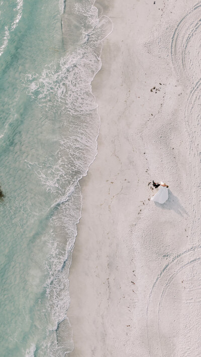 Aerial view of bride and groom walking on the beach near ocean waves during their wedding day.