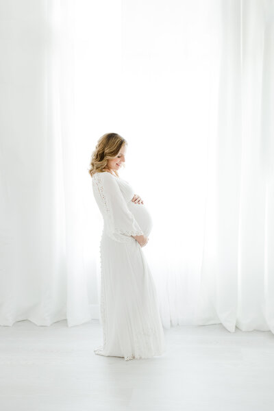 Pregnant woman wears long flowing white dress and smiles while holding her belly during maternity portrait session