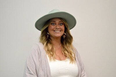 A portrait of Marie Cooper, a stylist at 212 Salon, Spa, & Barbershop, wearing a wide-brimmed hat and smiling at the camera.