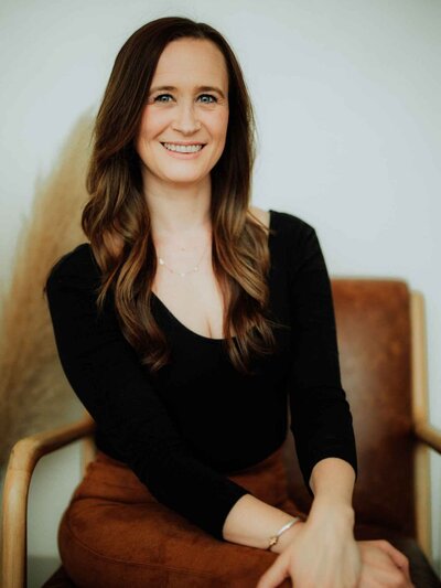 A woman with long blonde hair, wearing a black long-sleeve shirt and pants, sits on a tan chair next to dried pampas grass, smiling at the camera in a bright, minimalist room at Falmouth Maine Medical Aesthetics.