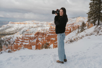 Woman holding her sony while looking at the camera smiling standing in snow. Bryce Canyon rocks covered in snow in the back ground with its orange peaks peeking through 