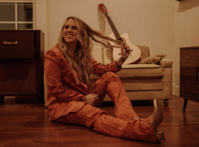 The Frisco hair color specialist is sitting on the floor and is wearing a dark orange outfit. Behind her is a beige sofa with an acoustic guitar and she is holding a part of her long wavy hair.