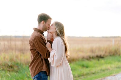 A couple embracing outdoors, with the man gently leaning his forehead against the woman's. The woman is wearing a navy off-the-shoulder dress and smiling.