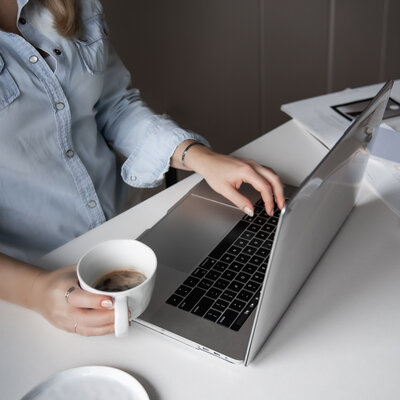 Close-up of a woman in a light blue shirt working on a laptop while holding a cup of coffee, representing focus and productivity, ideal for showcasing streamlined business systems and efficient workflows