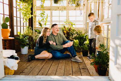 Family mini session photo in a beautiful spot with green plants and wooden floor. The parents are sitting with green outfits and the kids are in front of a window with sunlight