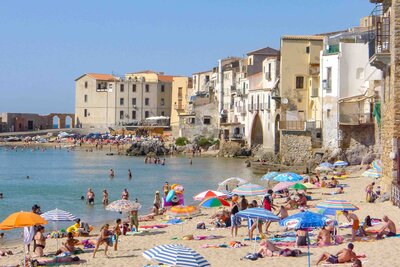 Sandy beach in Cefalù, Sicily, Italy, filled with colorful umbrellas and sunbathers, with turquoise waters and historic seaside buildings lining the shore ©Stephanie Dosch | theViatrix Luxury Travel