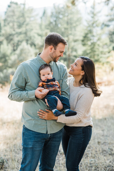 outdoor family pictures with denver family photographers capturing mother and father embracing as they hold their baby together in the woods
