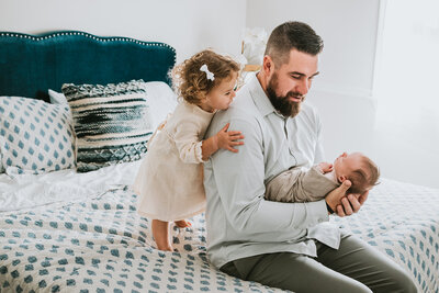 Dad holding newborn baby girl while looking at her and sitting on a bed while sister is standing behind dad looking at newborn baby in Davidsonville Maryland.