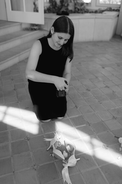 Photographer Hallie in an elegant black dress focuses on adjusting her camera in a sunlit New York City street, the light creating dramatic shadows around her, capturing her dedication to her craft, by Hallie Noel Photography.