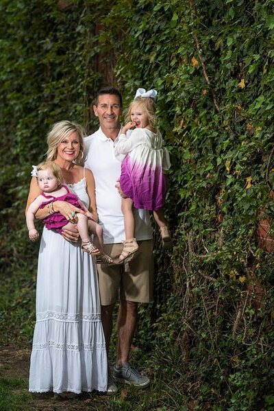 A family poses happily against a lush green ivy wall. The mother holds a baby in a magenta dress, while the father holds a child wearing a white and purple dress.