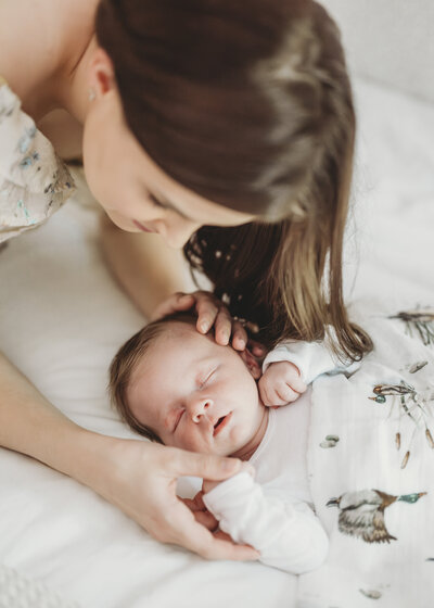 Mother smiling down at her newborn baby lying on a soft white bed during a lifestyle newborn session in Kansas City.