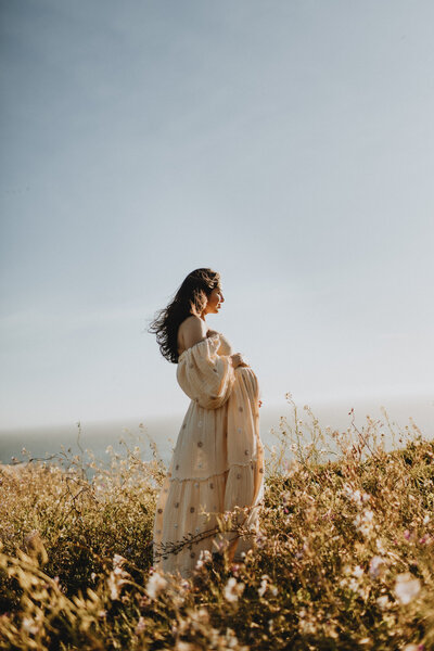 A woman in a flowing dress stands in a field of wildflowers, looking out at the ocean under a clear blue sky. her hair blows gently in the breeze, capturing a serene moment during maternity photoshoot.