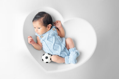 Newborn baby in a light blue outfit laying on a white backdrop with a heart outline. 