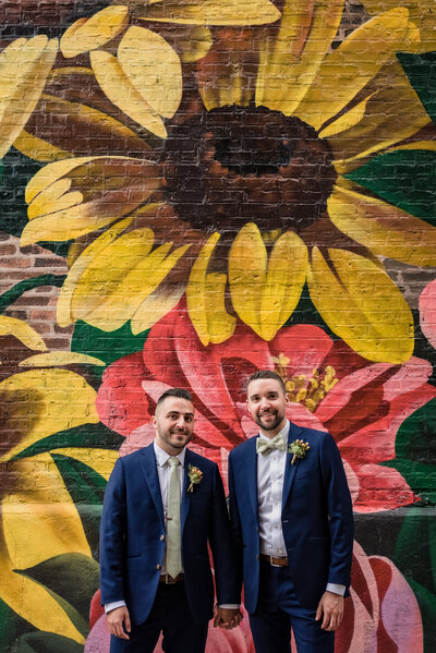 Two grooms hold hands in front of flower mural at Homestead on the Roof in Chicago, IL