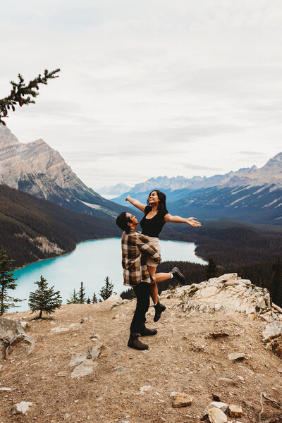 This is a picture of a couple  standing on top of a mountain. The husband is holding up  his new wife spinning her around. They are so happy, it is their elopement day. They can see a pretty blue lake down in the valley.
