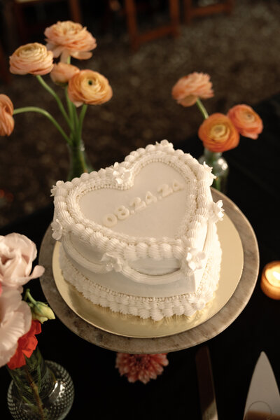 A heart-shaped white wedding cake with intricate piping details and small floral decorations sits on a marble stand. The date "08.24.24" is embossed on top. Fresh flowers and candles surround the cake.