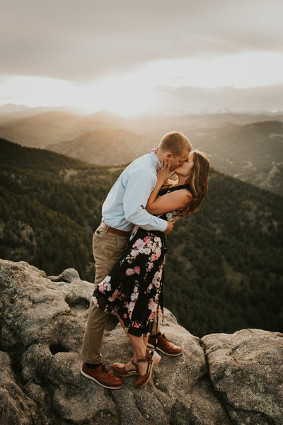 man and woman standing on  cliff kissing with mountains in  background