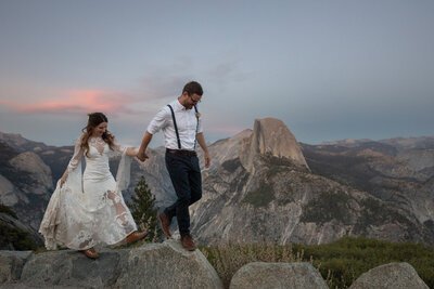 A couple walks hand in hand across rocky terrain during their Yosemite elopement at Glacier Point, with Half Dome in the background and a soft pink sunset sky above. Captured by Yosemite elopement photographers, this adventurous moment showcases the beauty of an intimate wedding in the park.