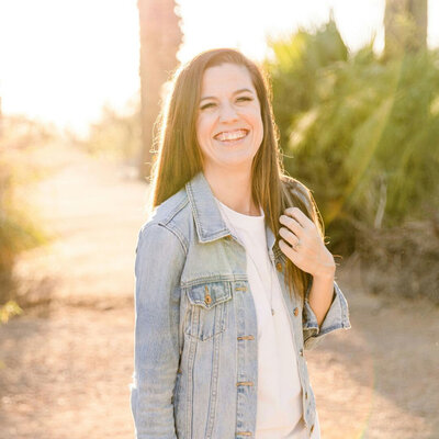 A woman with long brown hair is standing outdoors, smiling brightly. She is wearing a light blue denim jacket over a white top, and the background features a sunlit path with palm trees.