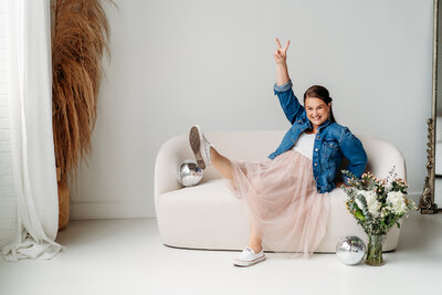 Female in jean jacket and light pink skirt kicking leg up on a couch.