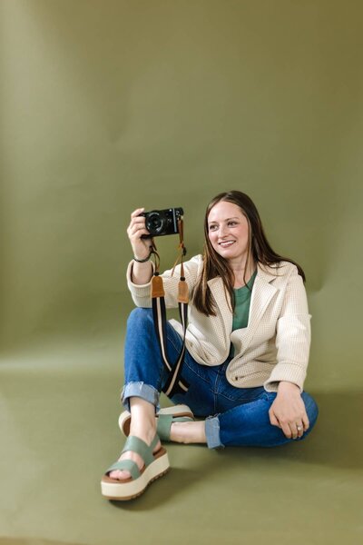 McKenna Fahrenbruch, content creator and co-founder of Graceful Exposure Media, sitting in front of a green backdrop holding her professional camera, ready for a branding photography session.