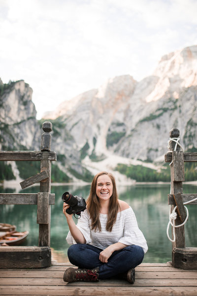 Photographer, Aris Ringas, sits on a wooden deck holding her camera. The deck overlooks a teal colored lake with mountains rising behind it.