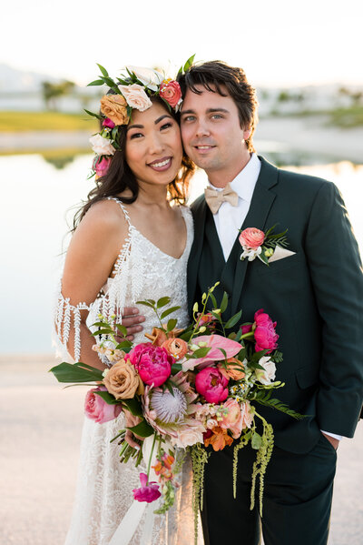A bride adorned with a fresh floral headband with a matching bouquet, standing cheek to cheek with her husband in front of a pond at Bali Hai Golf Club in Las Vegas, Nevada. 