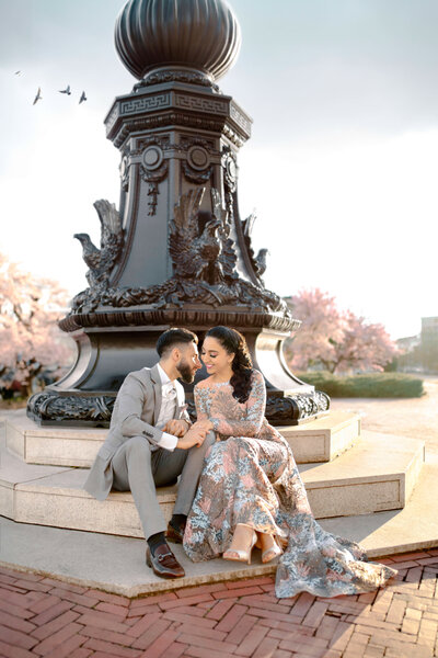 A couple sits lovingly in front of an ornate black lamp post with bird sculptures. The man wears a light gray suit, and the woman is in a floral dress. Cherry blossoms and a few birds in flight are seen in the background.